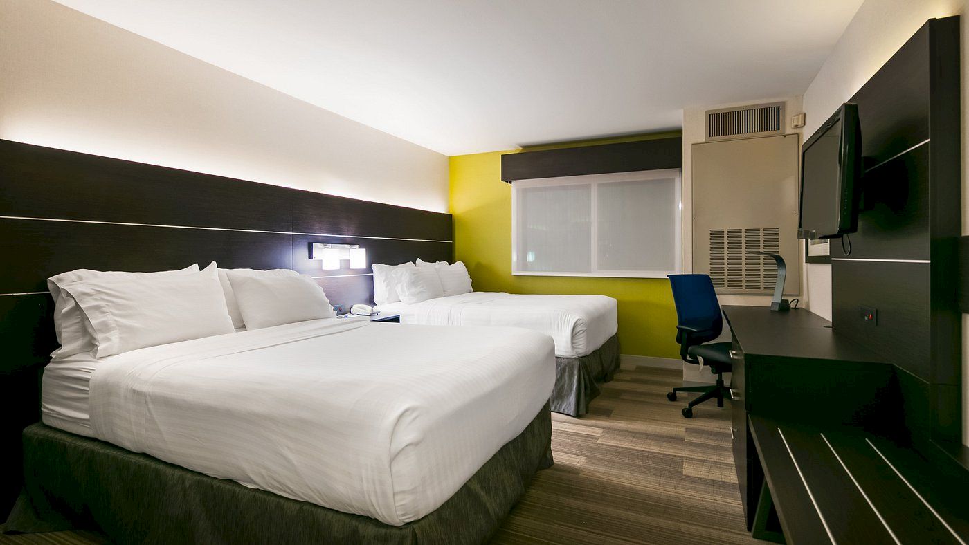 A hotel room with two double beds, white linens, nightstands, a desk with a chair, a flatscreen TV, and modern decor with a yellow accent wall.