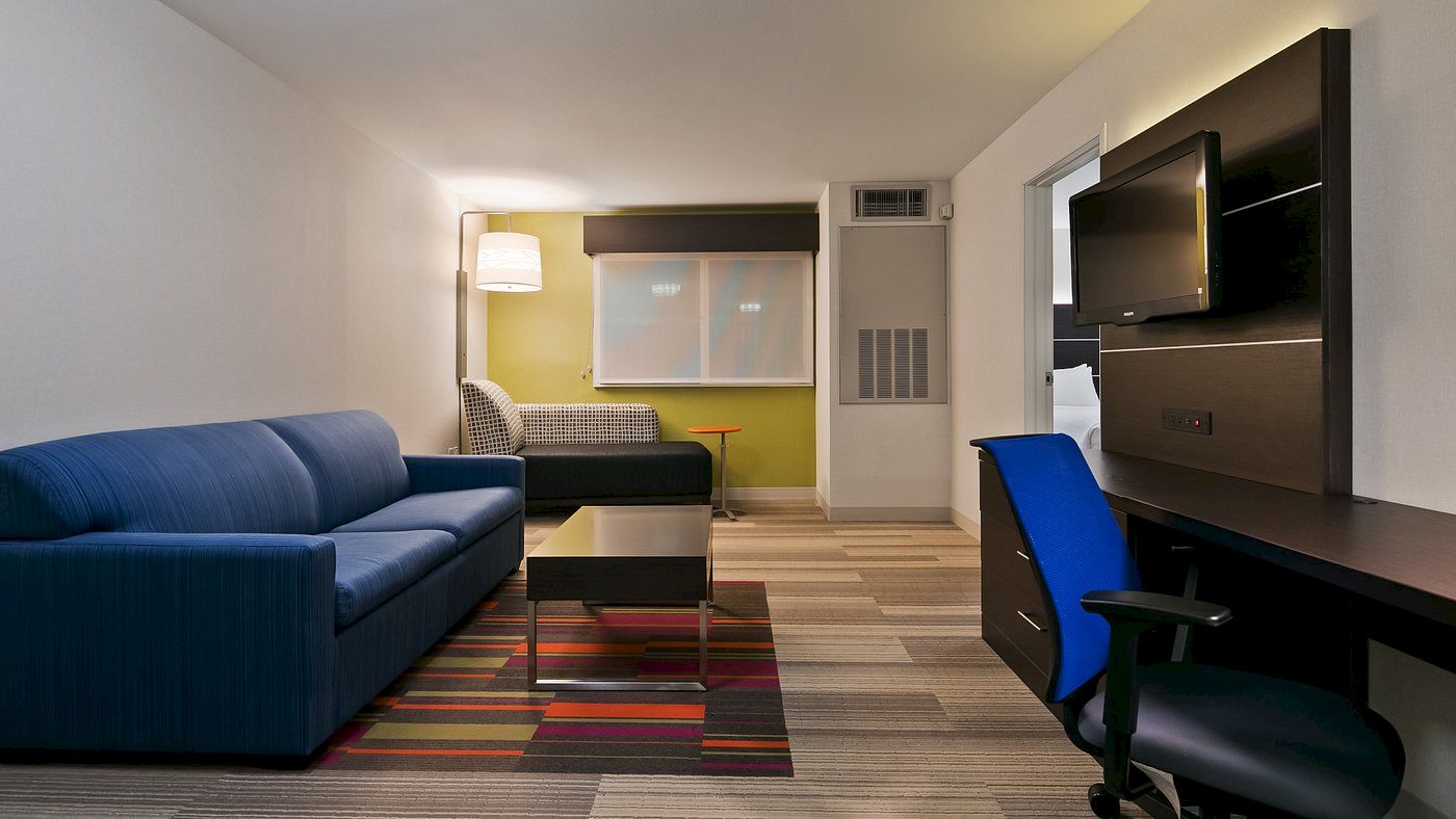 A modern living room with a blue sofa, a flat-screen TV, a desk with a blue chair, a table, and a lamp, all set on a striped carpet.