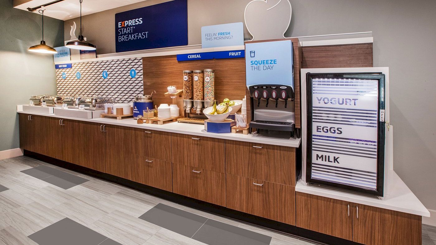 A hotel breakfast buffet with cereal, fruit, yogurt, eggs, and milk. Signage includes "Express Start Breakfast" and "Squeeze The Day".