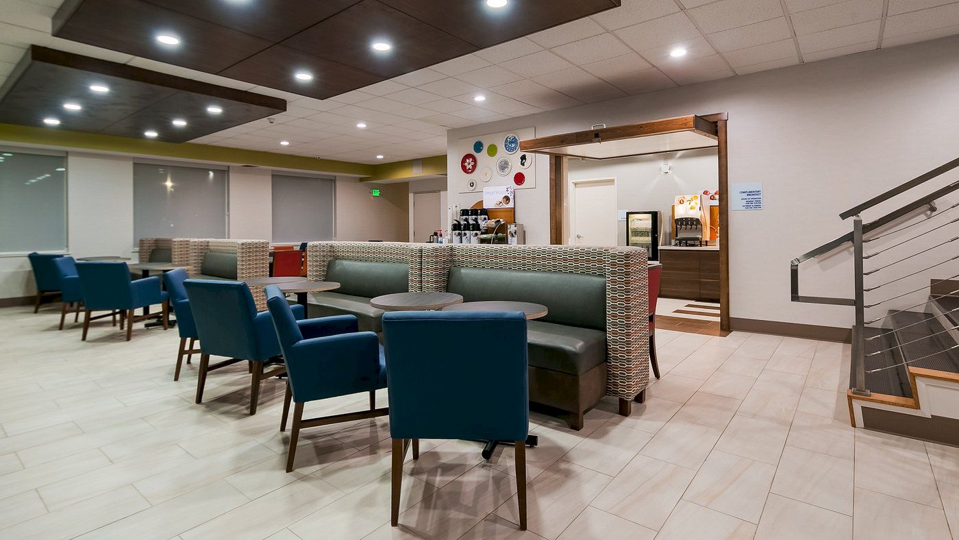 The image shows a modern, clean dining area with blue and green seating, tables, a beverage station in the back, and a staircase to the side.