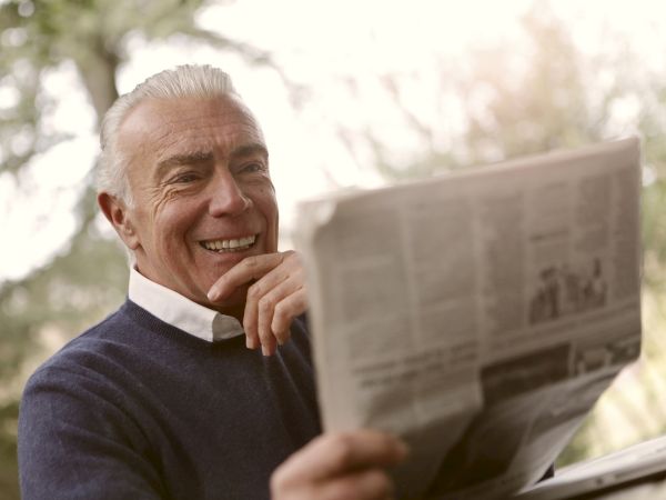 A person is smiling while reading a newspaper outdoors, dressed in a dark sweater and white shirt, with trees blurred in the background.