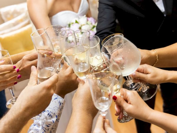 People toasting with champagne glasses at a celebration, likely a wedding, given the attire with a bride in a white dress holding a bouquet.