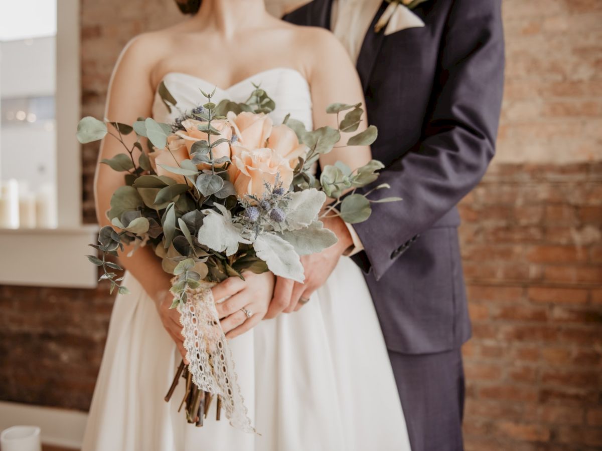 A couple dressed in wedding attire stands together, with the focus on the bride holding a bouquet of flowers while being embraced.