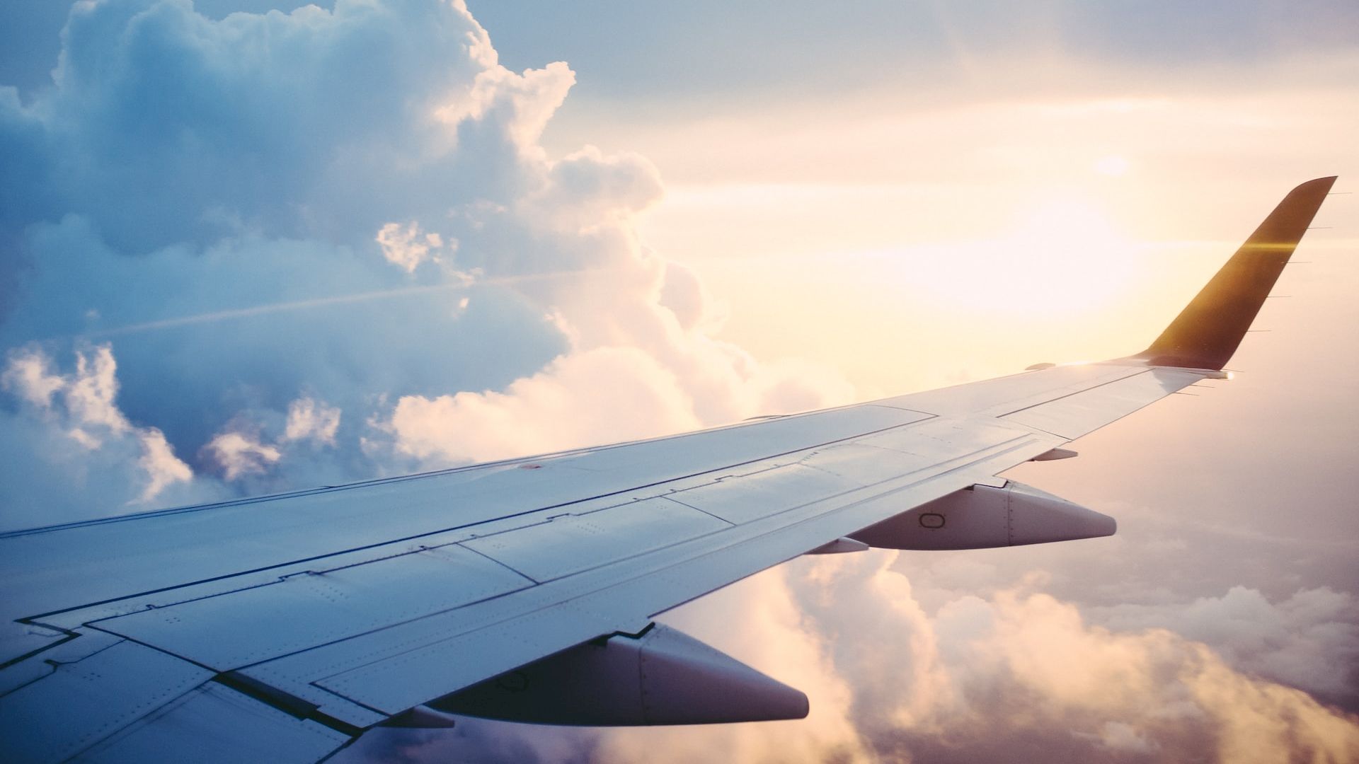The image shows an airplane wing with sunlight in the background, surrounded by clouds, giving a serene view from the window of the aircraft.