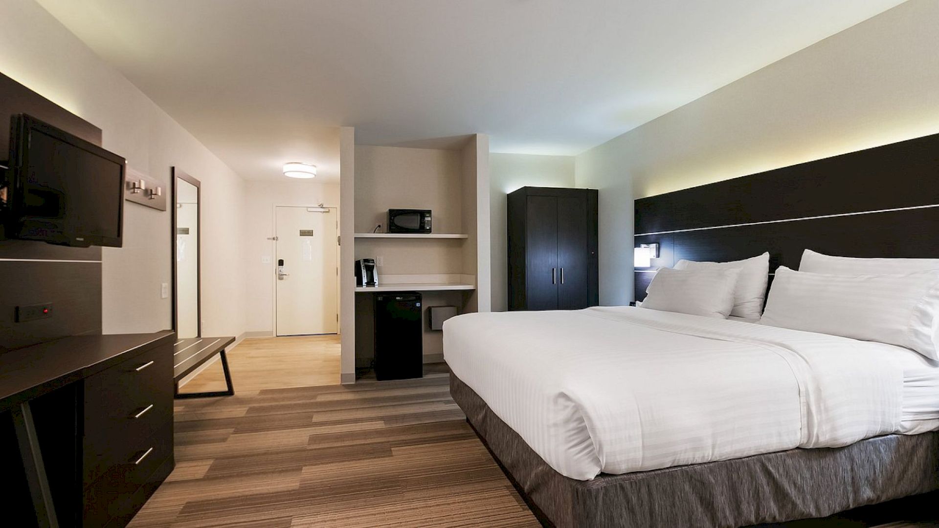 A modern hotel room with a large bed, flat-screen TV, wardrobe, desk, and a microwave on a shelf. The room has wood flooring and neutral decor.