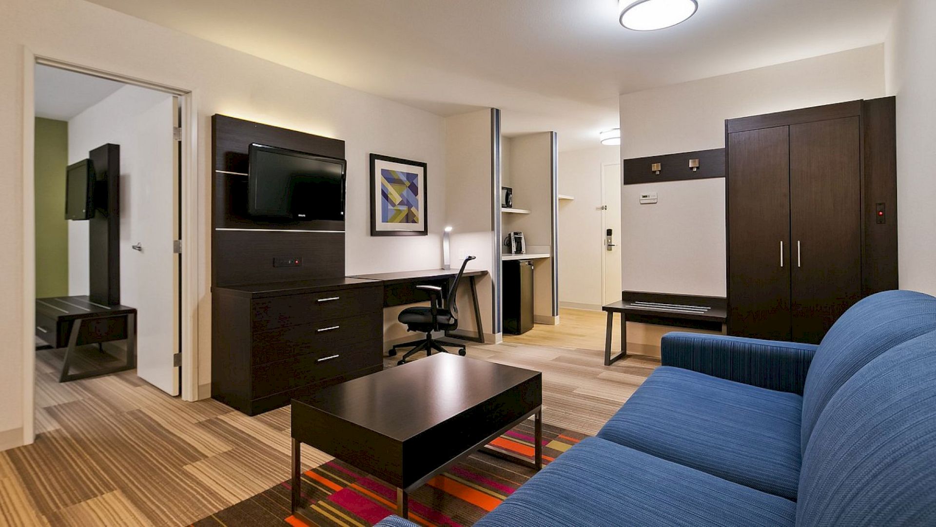 A modern hotel suite with a blue sofa, desk, flat-screen TV, and wall art. It has a bedroom, work area, and a small hallway.