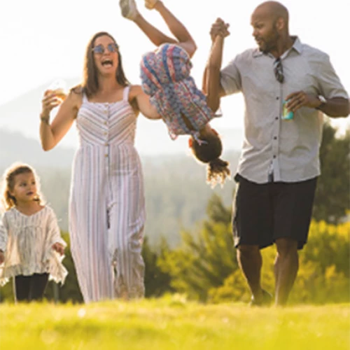 A family of four is outdoors, with two adults swinging a child upside-down between them while another child runs beside them on a sunny day.