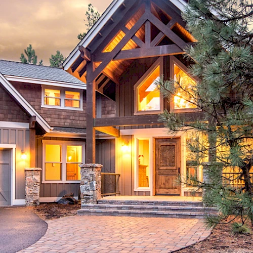 A beautiful rustic house with warm lighting, surrounded by trees, featuring a stone-paved pathway, a garage, and large windows at dusk.