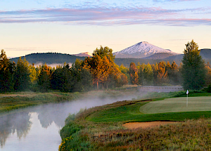 A picturesque golf course with a pathway near a river, trees, and a snow-capped mountain in the background under a colorful sky at dawn or dusk.