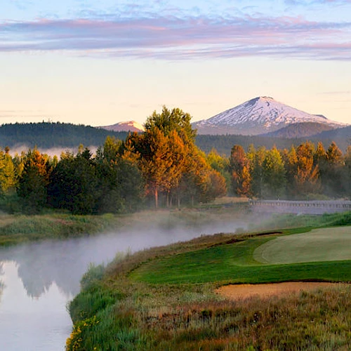 A picturesque golf course with a pathway near a river, trees, and a snow-capped mountain in the background under a colorful sky at dawn or dusk.