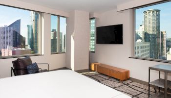 A modern hotel room with a bed, armchair, wall-mounted TV, and large windows offering a city skyline view.