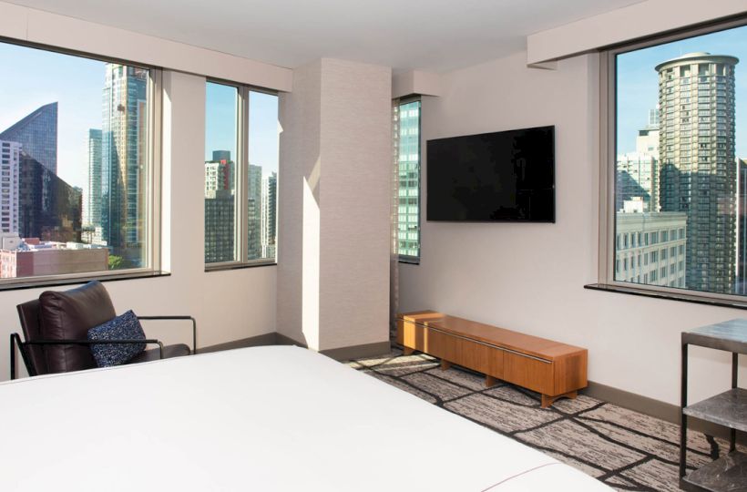 A modern hotel room with a bed, armchair, wall-mounted TV, and large windows offering a city skyline view.
