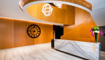 A modern lobby with a marble front desk, wooden walls, a large black clock, spiral stairway, hanging lights, and a floral arrangement on the counter.