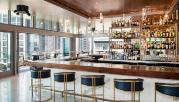 Modern bar with a wooden countertop, five blue and gold stools, and a fully stocked liquor display, featuring large windows and city views.
