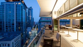 The image depicts a modern balcony at dusk, overlooking a cityscape with tall buildings and a street below. The balcony has tables and chairs.