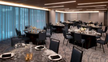 The image shows an elegant banquet hall with round tables set for dining, featuring black napkins, white flowers, and dim lighting, ready for an event.