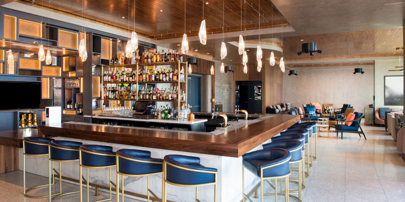 A modern bar with blue stools, a well-stocked liquor shelf, hanging lights, and a cozy seating area in the background.