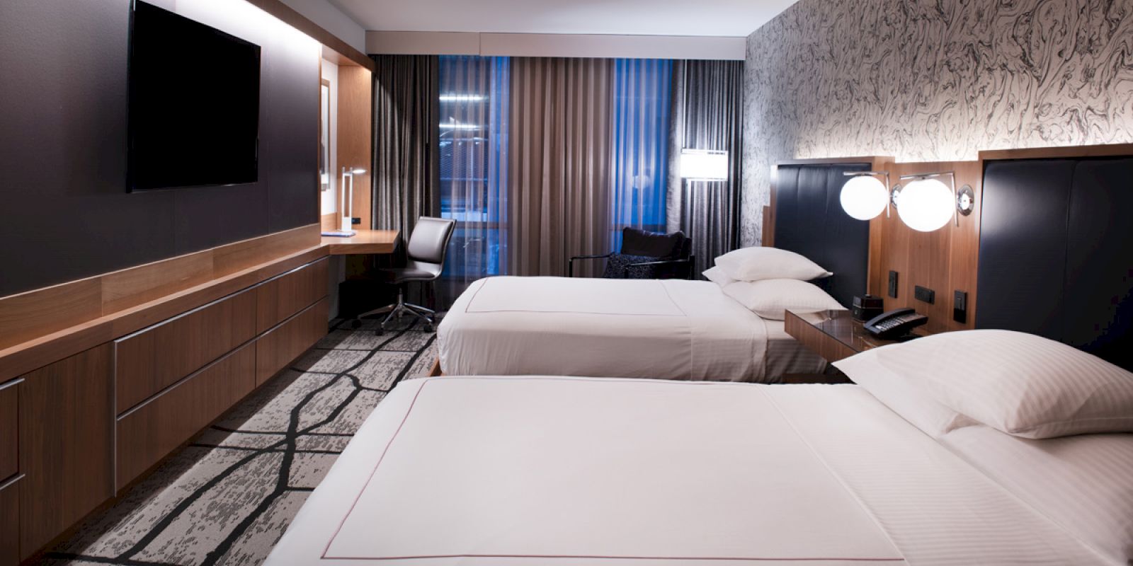 The image shows a modern hotel room with two neatly made beds, a wall-mounted TV, a desk with a chair, and large windows with curtains.