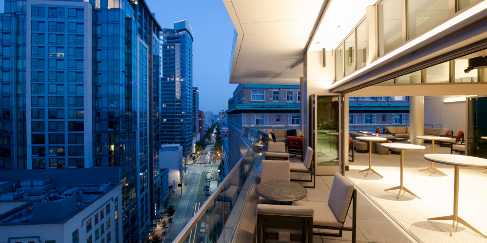 The image shows a modern balcony with outdoor seating, overlooking a cityscape of illuminated high-rise buildings at dusk.