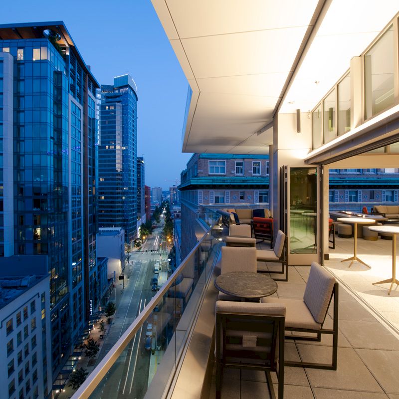 A modern cityscape at dusk, with tall buildings and a view from a stylish, illuminated balcony featuring outdoor seating and tables.