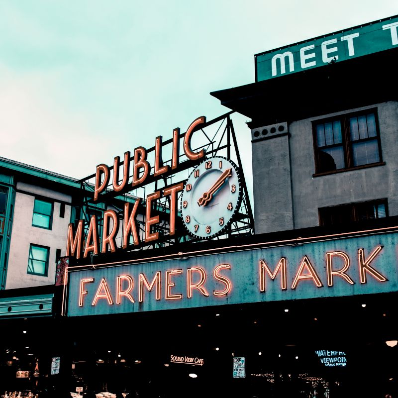 The image shows the exterior of a public market with neon signs for "Public Market" and "Farmers Market," and buildings surrounding the area.