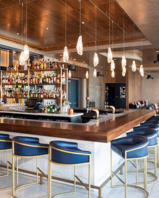 The image shows a modern bar with stylish lighting, a stocked liquor shelf, and blue bar stools. There is a seating area visible in the background.