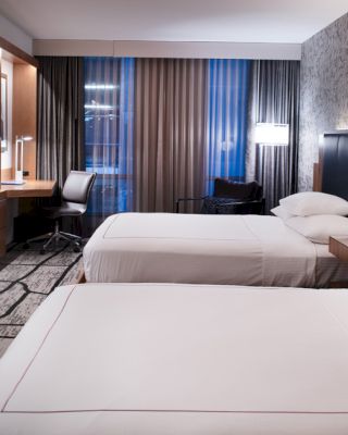 A modern hotel room features two neatly made beds, a wall-mounted TV, a desk with a chair, and contemporary decor with soft lighting.