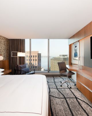 A modern hotel room with a large bed, desk, chair, flat-screen TV, and a window showcasing a cityscape and water view in the background.