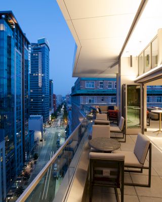 A modern cityscape view from a high-rise balcony with seating and tables, overlooking buildings and a street at dusk with building lights on.
