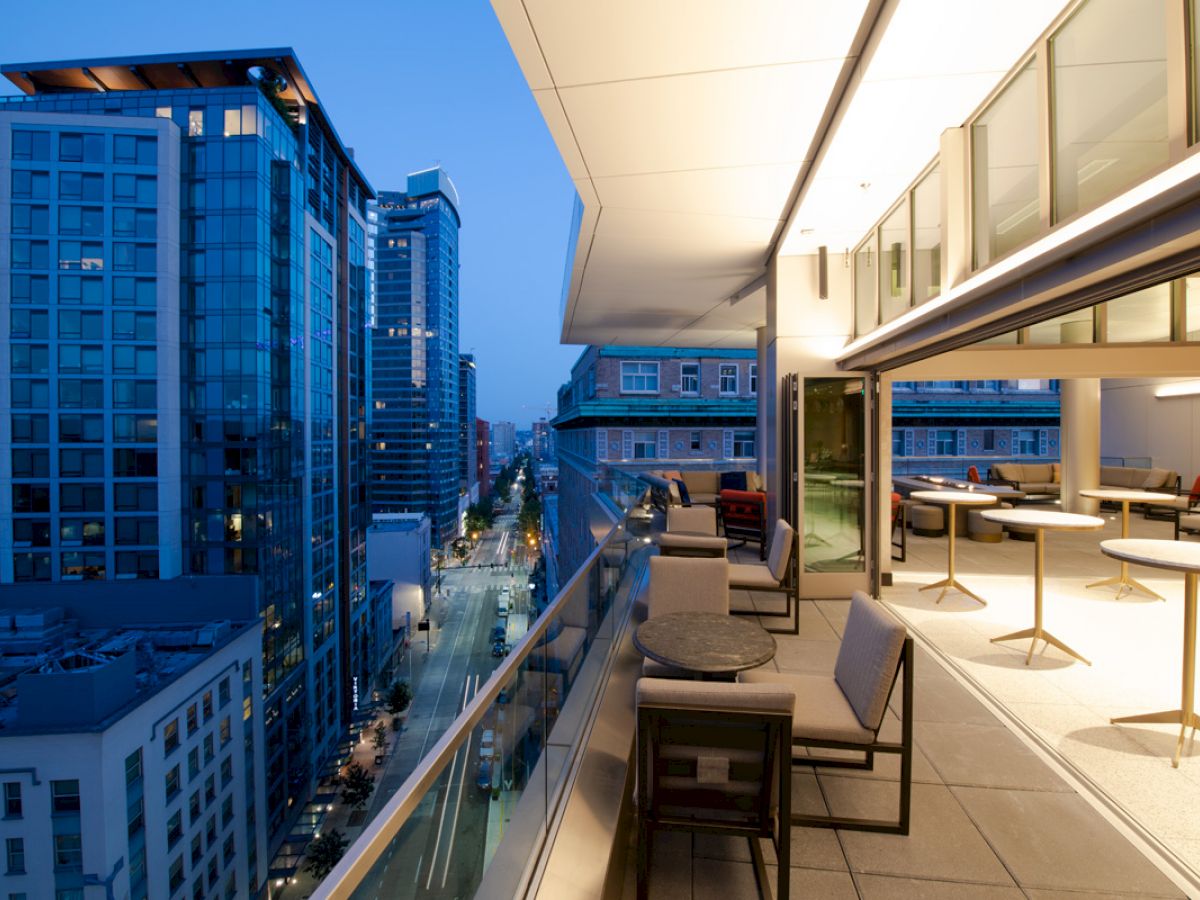 A modern cityscape features tall glass buildings and a street view, with an adjacent balcony hosting tables and chairs for outdoor seating.