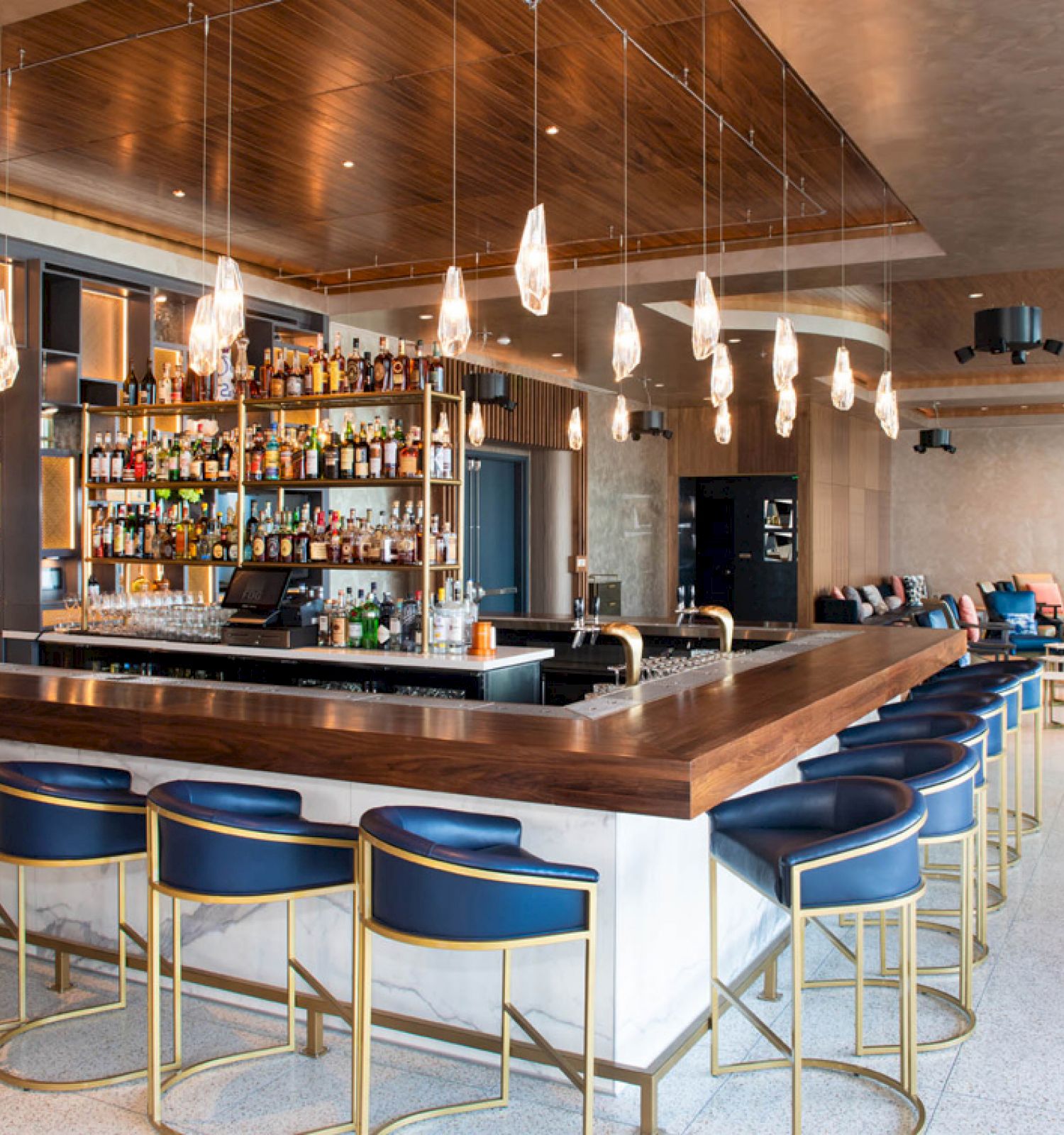 A stylish bar with a large selection of bottles, modern seating, hanging lights, and a lounge area in the background.