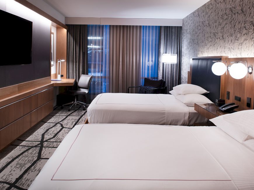 The image shows a hotel room with two neatly made beds, a wall-mounted TV, a desk with a chair, and modern decor including a patterned wallpaper.