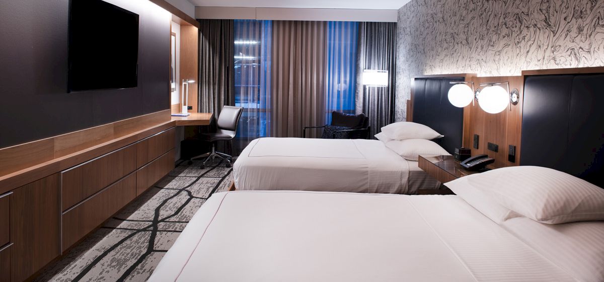 The image shows a hotel room with two neatly made beds, a wall-mounted TV, a desk with a chair, and modern decor including a patterned wallpaper.