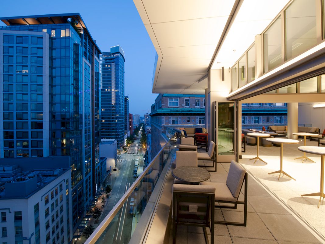 A rooftop terrace overlooks a city street, featuring modern outdoor furniture, glass railings, and adjacent tall buildings illuminated during dusk.