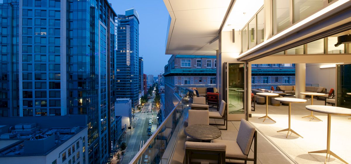 A rooftop terrace overlooks a city street, featuring modern outdoor furniture, glass railings, and adjacent tall buildings illuminated during dusk.