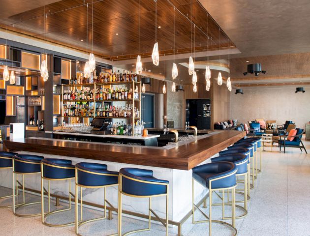 A modern bar interior with a well-stocked liquor shelf, hanging pendant lights, and blue barstools, surrounded by a spacious seating area.
