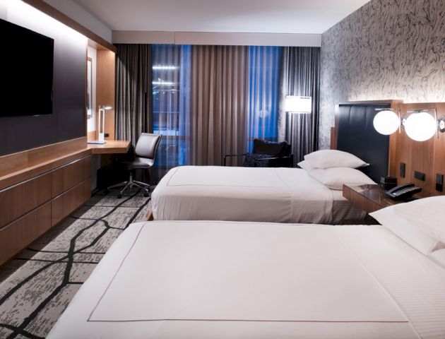 A modern hotel room with two beds, a wall-mounted TV, a desk with a lamp, and large windows with curtains and a patterned accent wall.