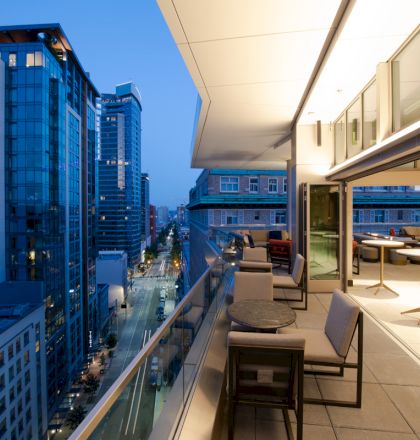 A cityscape view from a modern balcony at dusk, featuring outdoor seating and tables, with tall buildings and a street below.