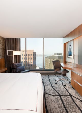 A modern hotel room with a large bed, desk with a chair, a wall-mounted TV, and a window view of city buildings and water.