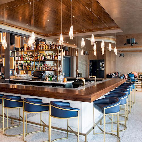 The image depicts a modern bar with a wooden counter, stylish bar stools, overhead lighting, and shelves stocked with various beverages.