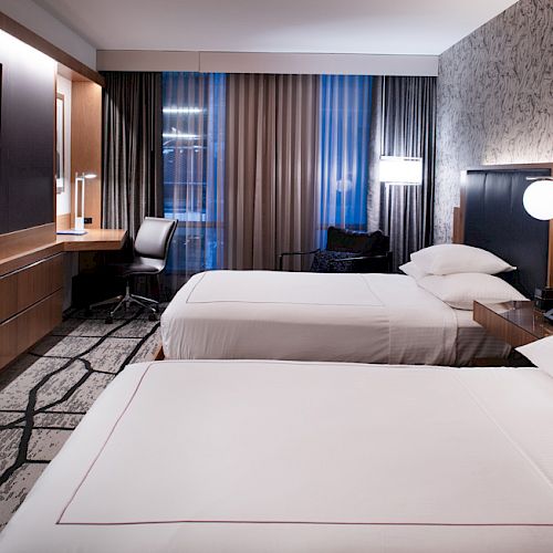 This image shows a modern hotel room with two beds, a flatscreen TV on the wall, a desk with a chair, and a stylish decor.