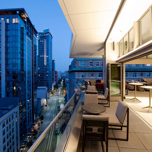 A modern cityscape at dusk with high-rise buildings and a spacious, well-lit balcony featuring seating arrangements and tables.