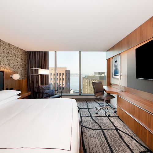 A modern hotel room features a large bed, a desk with a chair, a flat-screen TV, and a window view of buildings and water in the background.