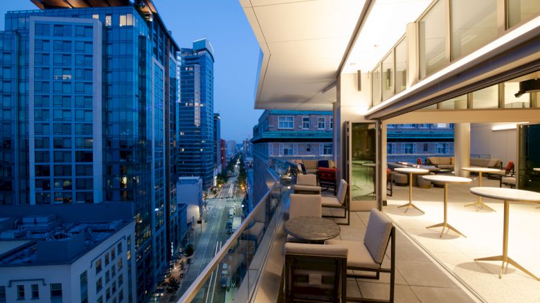 A modern cityscape with tall buildings and a cozy, well-lit balcony featuring tables and chairs overlooking the bustling street below.