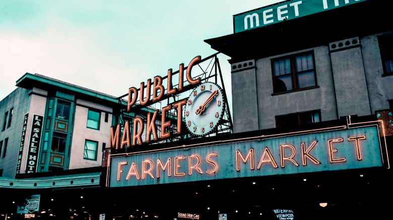 The image shows a neon sign for a "Public Market" alongside a "Farmers Market" sign, and surrounding buildings, likely at an iconic market location.