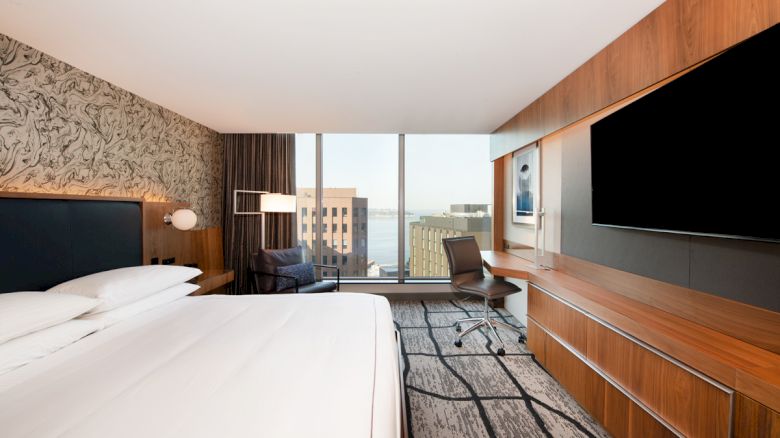 The image shows a modern hotel room with a large bed, wall-mounted TV, desk, chair, and window offering a city and ocean view.