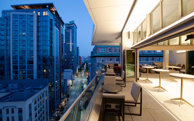 A modern cityscape at dusk with a view from a high-rise balcony, featuring outdoor seating and adjacent buildings.