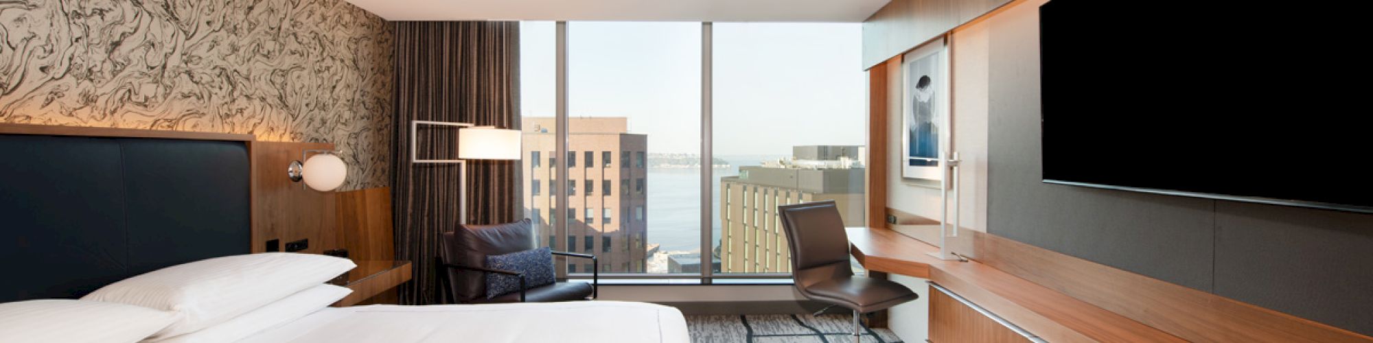 A modern hotel room with a large bed, desk, chair, television, and window with a city and water view in the background.